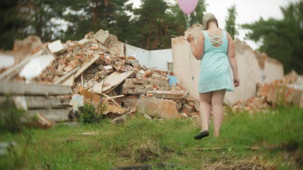 A little girl in a gas mask walks through the ruined buildings with balloons in her hand — Stock Video