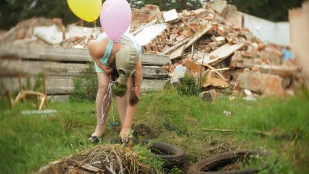 A little girl in a gas mask walks through the ruined buildings with balloons in her hand — Stock Video