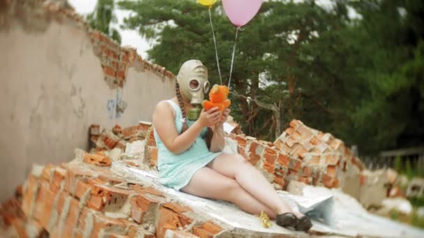 Een klein meisje in een gas mask op de ruïnes van een gebouw en vasthouden aan een pop en ballonnen. — Stockvideo