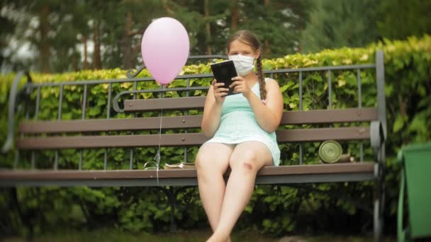 Linda chica en un respirador utiliza tableta en el parque en un banco — Vídeos de Stock