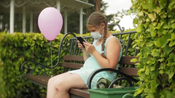 Roztomilá dívka v respirátor používá tabletu v parku na lavičce — Stock video