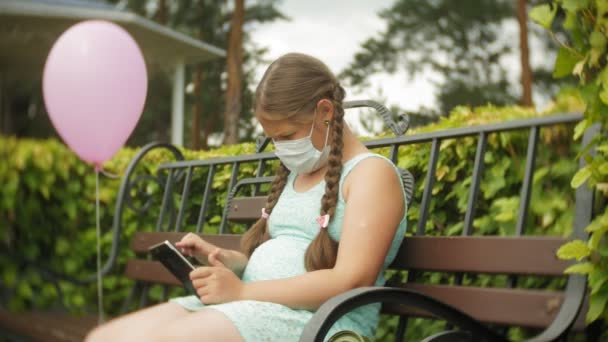 Linda chica en un respirador utiliza tableta en el parque en un banco — Vídeo de stock