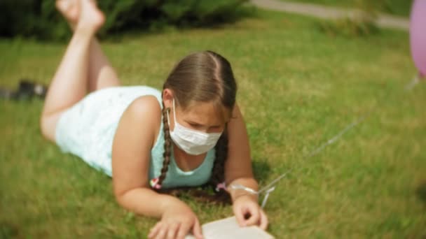 Nettes Mädchen mit Atemschutzmaske liest im Park ein Buch — Stockvideo