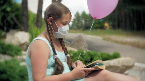 Nettes Mädchen mit Atemschutzmaske liest im Park ein Buch — Stockvideo
