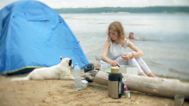 Femmes cuisiner des aliments dans bowler dans le camping avec tente sur le fond. le chien se promène à côté — Video