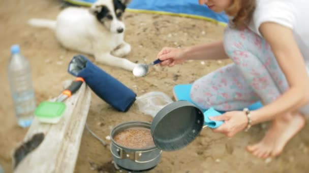 महिलाएं पृष्ठभूमि पर तम्बू के साथ शिविर में गेंदबाज में भोजन पका रही हैं। कुत्ता किनारे चलता है — स्टॉक वीडियो