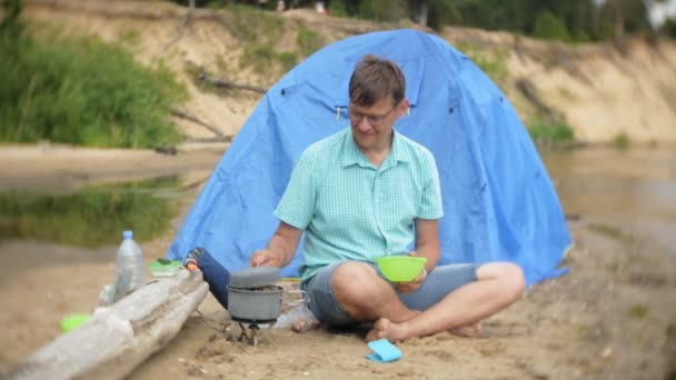 一个男人在一个露营地的水壶边吃饭, 背景上有帐篷。. — 图库视频影像