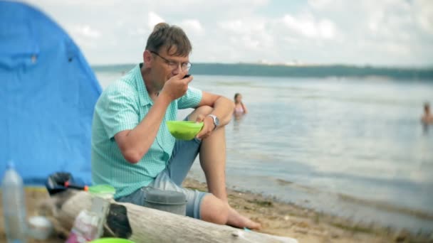 Een man is eten rond een waterkoker op een camping met een tent op de achtergrond. — Stockvideo