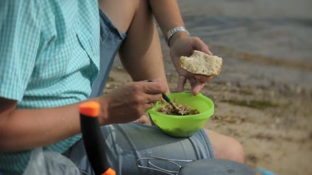 Een man is eten rond een waterkoker op een camping met een tent op de achtergrond. — Stockvideo
