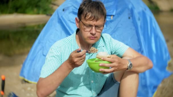 Ein Mann isst um einen Wasserkocher auf einem Campingplatz mit einem Zelt im Hintergrund. — Stockvideo