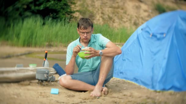 Un hombre está comiendo alrededor de una tetera en un campamento con una tienda de campaña en el fondo . — Vídeos de Stock