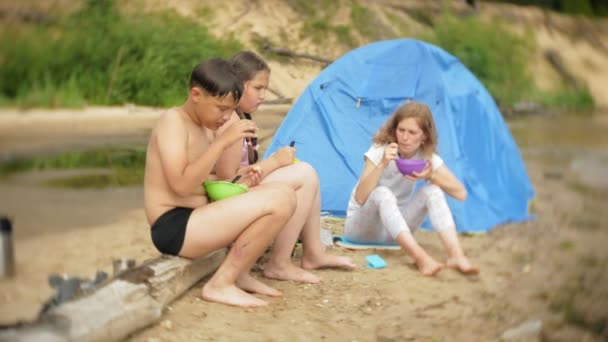 Eine Frau und ihre Kinder essen eine Mahlzeit in der Nähe des Wasserkochers auf einem Campingplatz mit einem Zelt im Hintergrund. — Stockvideo