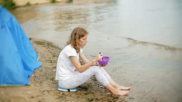 Een vrouw is het eten rond een waterkoker op een camping met een tent op de achtergrond. — Stockvideo