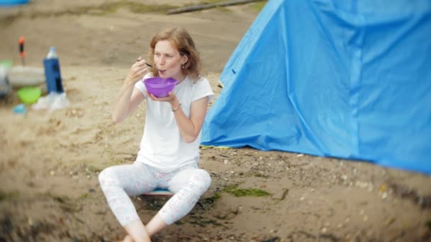 Een vrouw is het eten rond een waterkoker op een camping met een tent op de achtergrond. — Stockvideo