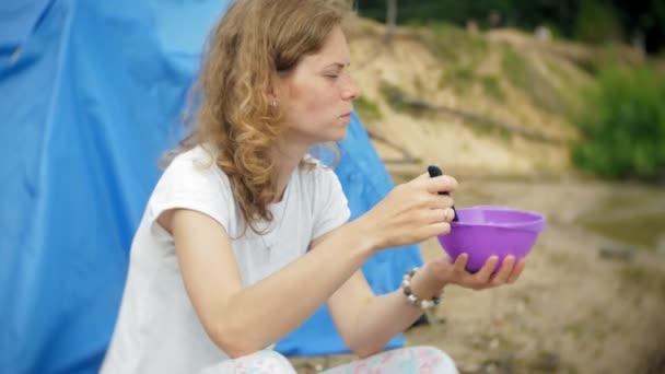 Una mujer está comiendo alrededor de una tetera en un campamento con una tienda de campaña en el fondo . — Vídeos de Stock