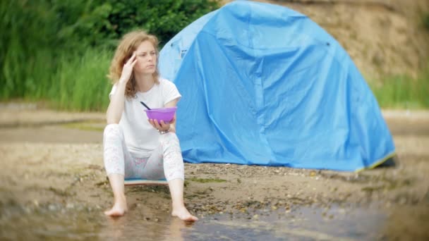 Eine Frau isst um einen Wasserkocher auf einem Campingplatz mit einem Zelt im Hintergrund. — Stockvideo