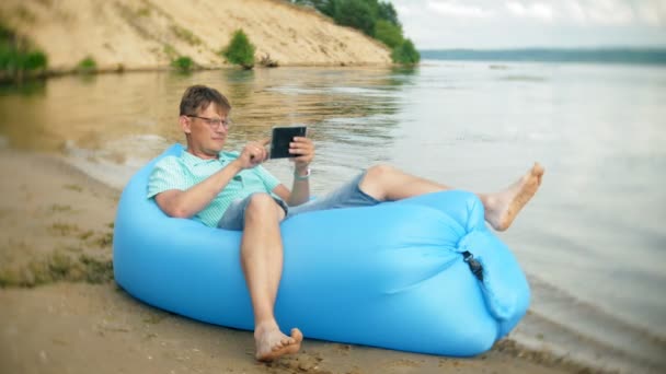 Мужчина отдыхает на надувном матрасе у моря. Он использует планшет — стоковое видео