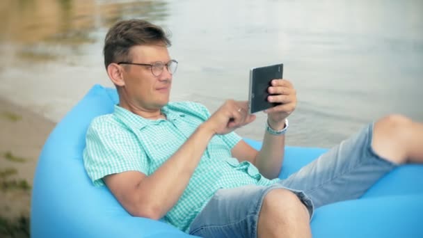 Чоловік відпочиває на надувному матраці біля моря. Він використовує планшет — стокове відео
