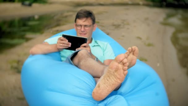 Un hombre está descansando en un colchón inflable junto al mar. Usa una tableta. — Vídeos de Stock