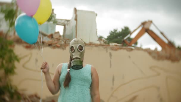 Маленькая девочка в противогазе ходит по разрушенным зданиям с воздушными шарами в руках — стоковое видео