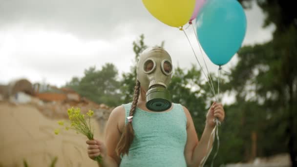 En liten flicka i en gasmask promenader genom de förstörda byggnaderna med ballonger i handen — Stockvideo