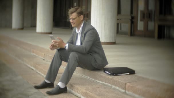 El hombre de negocios está sentado en las escaleras de la ciudad. Lleva un traje y un maletín y habla en el smartphone — Vídeo de stock