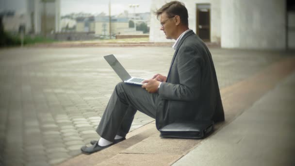 L'uomo d'affari e 'seduto sulle scale della citta'. Indossa un completo e una valigetta. Lavora su un portatile. — Video Stock