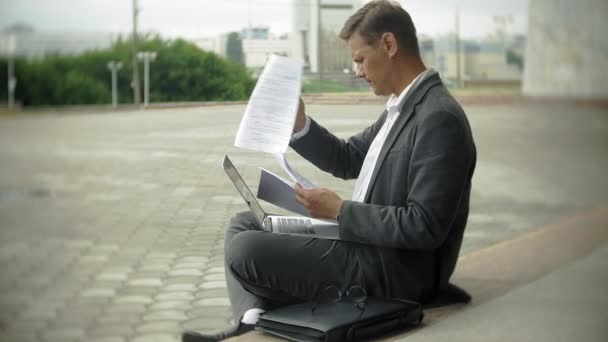 Businessman is sitting on the stairs in the city. He wears a suit and briefcase. He works on a laptop — Stock Video