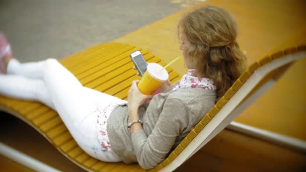 Mujer joven con batido y teléfono celular al aire libre en un cómodo banco creativo — Vídeos de Stock