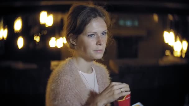 Молода жінка їсть картоплю фрі вночі на відкритому повітрі за допомогою світла ліхтарів — стокове відео