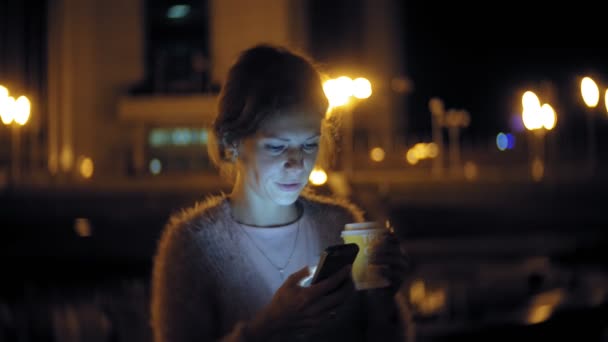 Mensajería de texto de mujer joven en el teléfono móvil mientras está en el centro por la noche. Mensajes femeninos felices con smartphone en la ciudad . — Vídeo de stock