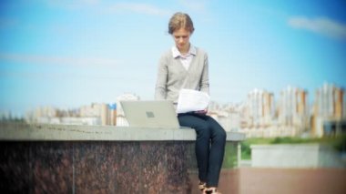 İş merkezinde bir dizüstü bilgisayar ile belgili tanımlık merdiven üzerinde oturan genç iş kadını belgeleri okuma.