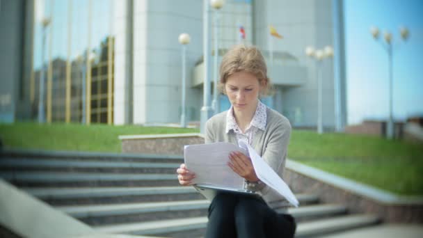 İş merkezinde bir dizüstü bilgisayar ile belgili tanımlık merdiven üzerinde oturan genç iş kadını belgeleri okuma. — Stok video
