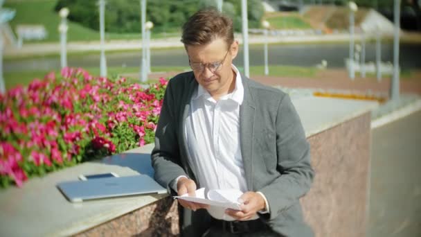 Young business man sitting on the steps with a laptop in the business center, reading documents. — Stock Video