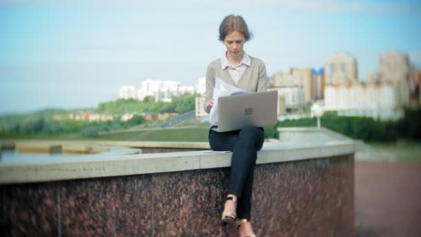 O tânără femeie de afaceri care stă pe trepte cu un laptop în centrul de afaceri, citind documente . — Videoclip de stoc