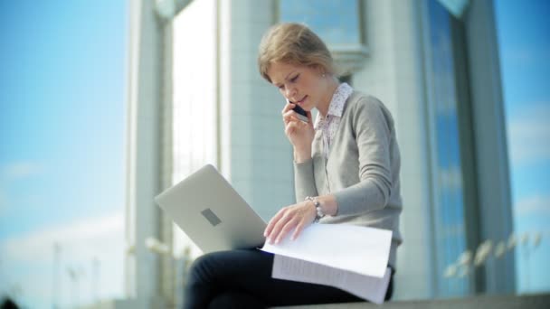 Jovem mulher de negócios sentada nos passos com um laptop no centro de negócios, lendo documentos . — Vídeo de Stock