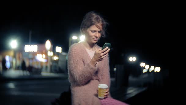 Mensajería de texto de mujer joven en el teléfono móvil mientras está en el centro por la noche. Mensajes femeninos felices con smartphone en la ciudad . — Vídeos de Stock