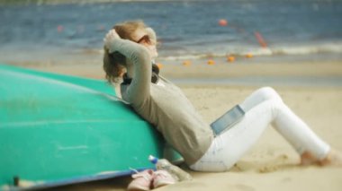 Genç güzel kadın kulaklık tekne yakınındaki kumsalda oturur ve bir tablet kullanır