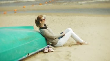 Genç güzel kadın kulaklık tekne yakınındaki kumsalda oturur ve bir tablet kullanır