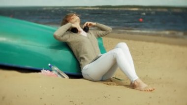 Genç güzel kadın kulaklık tekne yakınındaki kumsalda oturur ve bir tablet kullanır