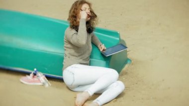 Genç güzel kadın kulaklık tekne yakınındaki kumsalda oturur ve bir tablet kullanır