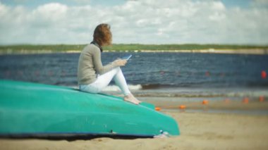 Genç güzel kadın kulaklık tekne yakınındaki kumsalda oturur ve bir tablet kullanır