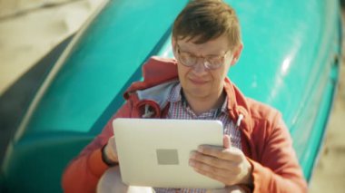 Yaşlı adam kulaklık tekne yakınındaki kumsalda oturur ve bir tablet kullanır