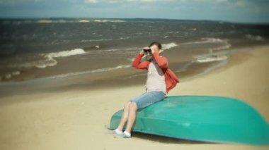 Yaşlı bir adam sahilde tekne dışında oturur ve dürbün ile görünüyor