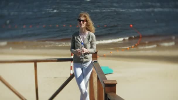 Женщина пьет воду из бутылки на пляже — стоковое видео
