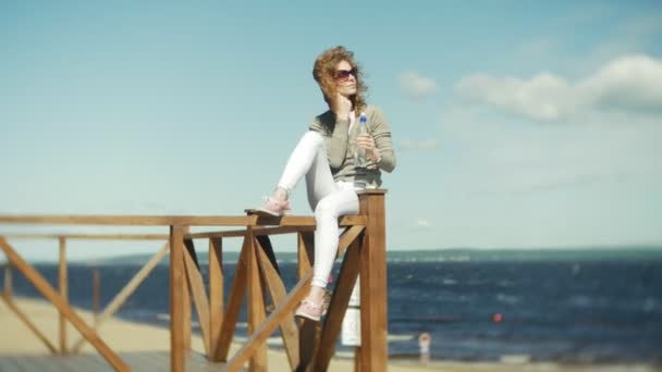 Mujer bebiendo agua de una botella en la playa — Vídeo de stock