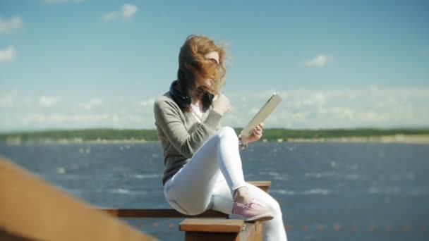 Joven hermosa mujer se sienta en la playa en los auriculares y utiliza una tableta — Vídeos de Stock