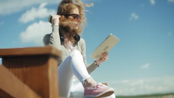 Joven hermosa mujer se sienta en la playa en los auriculares y utiliza una tableta — Vídeos de Stock