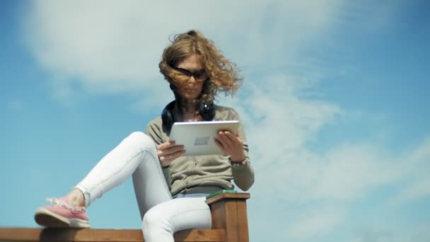 Giovane bella donna si siede sulla spiaggia in cuffia e utilizza un tablet — Video Stock