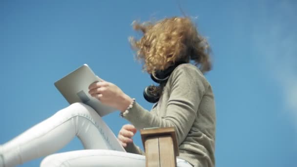 Νεαρή όμορφη γυναίκα κάθεται στην παραλία στα ακουστικά και χρησιμοποιεί ένα δισκίο — Αρχείο Βίντεο